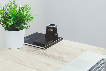 A computer desk with a green plant in a vase and a diary on the table.