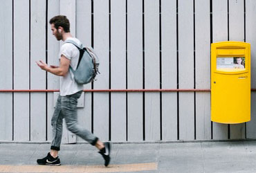 A man walking forward near the yellow postbox with a headset and phone in hand